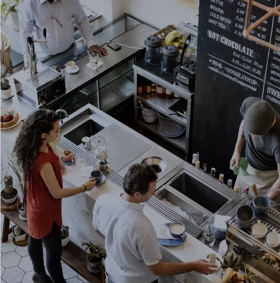 People at a cafe counter with a barista making drinks and a chalkboard menu in the background